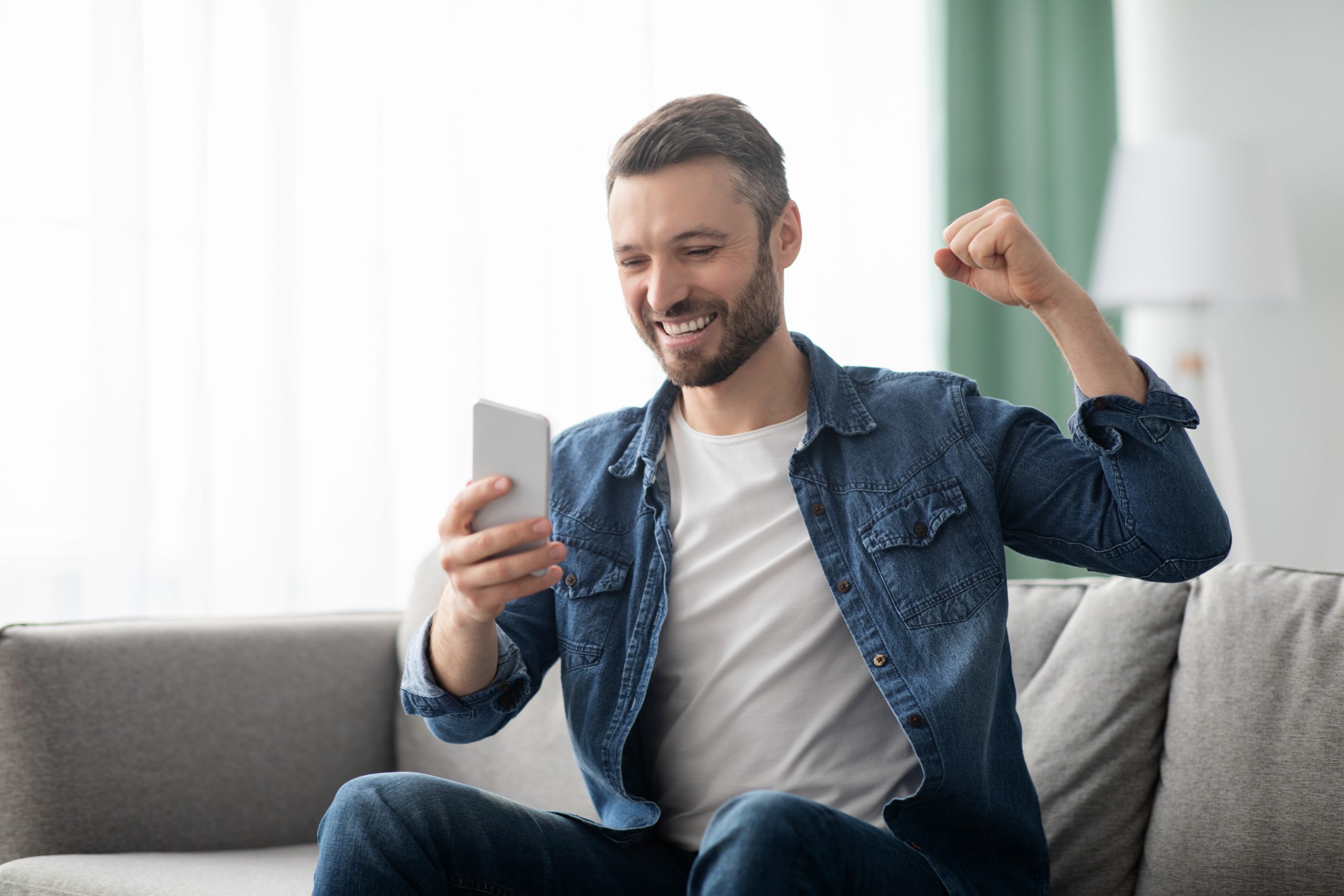 Emotional bearded middle-aged man celebrating success, gambling or trading online, using mobile phone while sitting on couch at home, copy space. Online gaming, financial markets concept